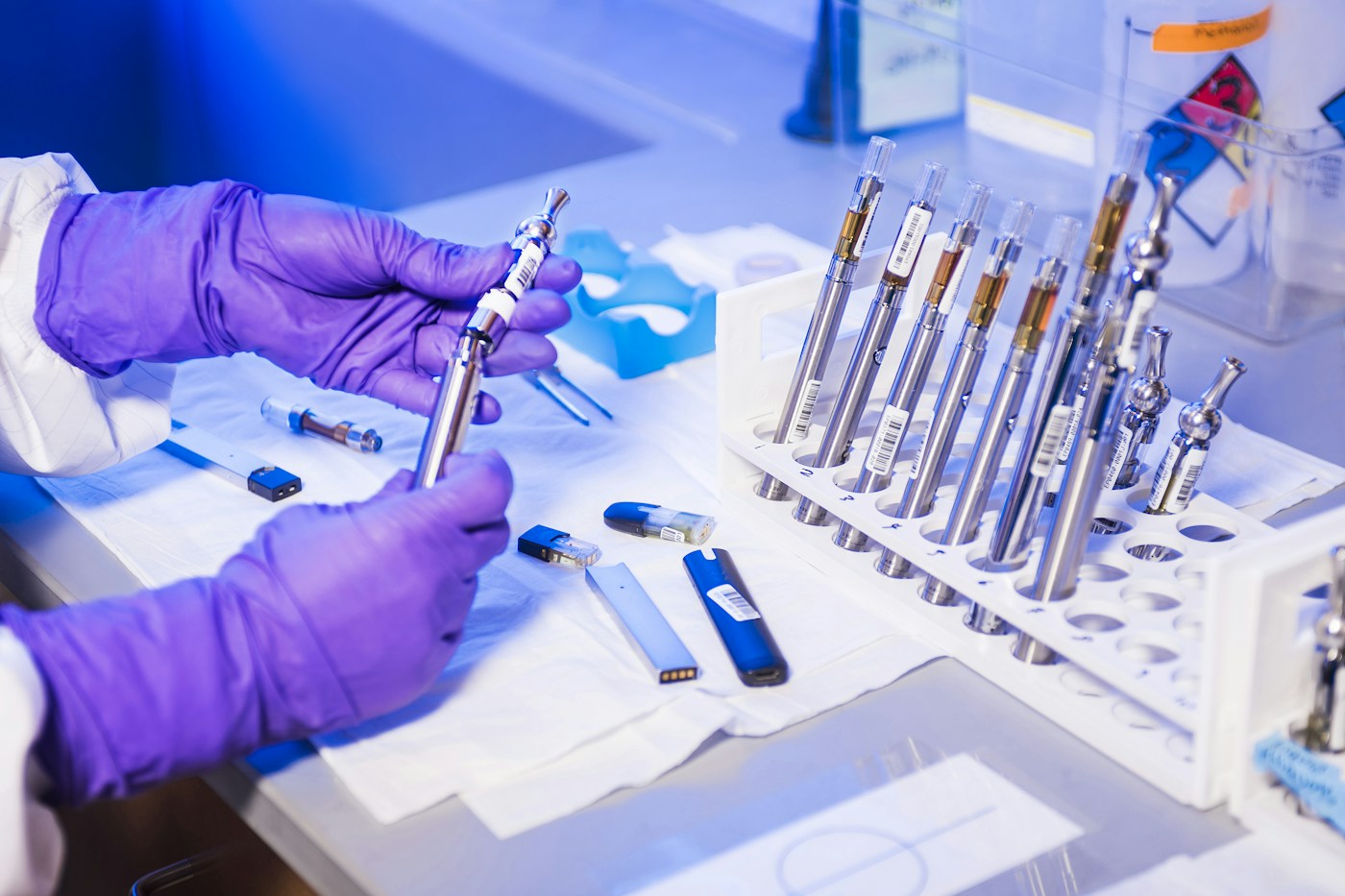Mani con guanti viola esaminano un dispositivo accanto ad altri campioni sul banco di laboratorio